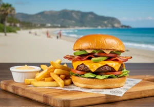 A Cali burger with a juicy patty, cheddar cheese, bacon, avocado, lettuce, and tomato on a wooden board with fries and dipping sauce.